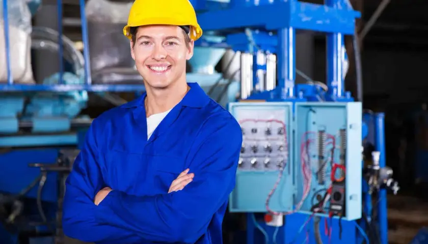 Uśmiechnięty pracownik w żółtym kasku i niebieskim kombinezonie, stojący przed panelem elektrycznym. To może być pracownik hurtowni elektrotechnicznej Comel Rumia.