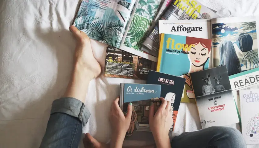 Osoba leży na łóżku otoczona **ciekawymi książkami po angielsku**. Widać fragmenty okładek z ilustracjami i tytułami.