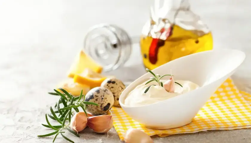 Kremowy sos aioli w białej miseczce, ozdobiony gałązką rozmarynu i ząbkiem czosnku. Obok składniki: jajka przepiórcze, cytryna, czosnek i oliwa.