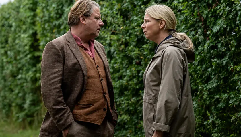 Två personer står vända mot varandra framför en grön häck. Mannen bär kavaj och väst, kvinnan en jacka. Scen från en dansk kriminalserie.