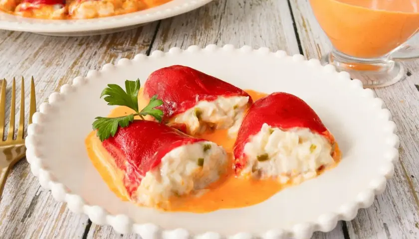 Pimientos rellenos de bacalao, cubiertos en una cremosa salsa naranja, servidos en un plato blanco con un tenedor dorado al lado.