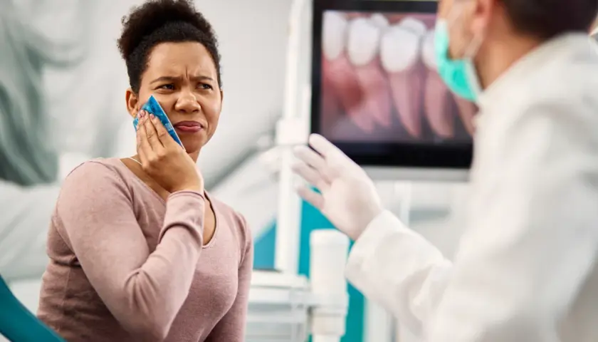 Mujer con compresa fría en la mejilla, sufriendo un alveolitis con dolor insoportable, mientras el dentista le explica algo.