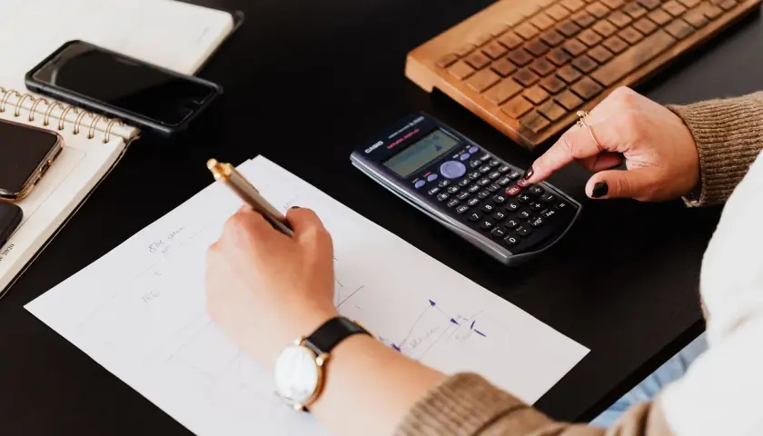 Mani che scrivono su fogli, usano una calcolatrice per lo scorporo IVA e una tastiera in legno.