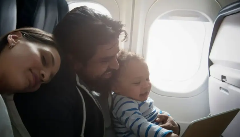 Familj flyger med bebis. Pappa håller ett leende barn som tittar på en surfplatta, mamman sover bredvid.