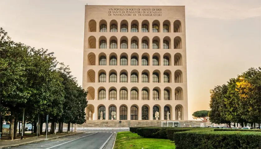 Edificio con arcate sovrapposte, simile a una stazione metro EUR Magliana, con iscrizione in latino e alberi verdi.