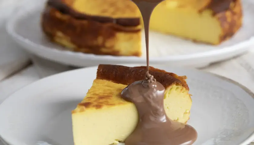 Un trozo de basque cheesecake cremoso recibe un chorro de chocolate fundido. Otro pastel entero se ve desenfocado al fondo.