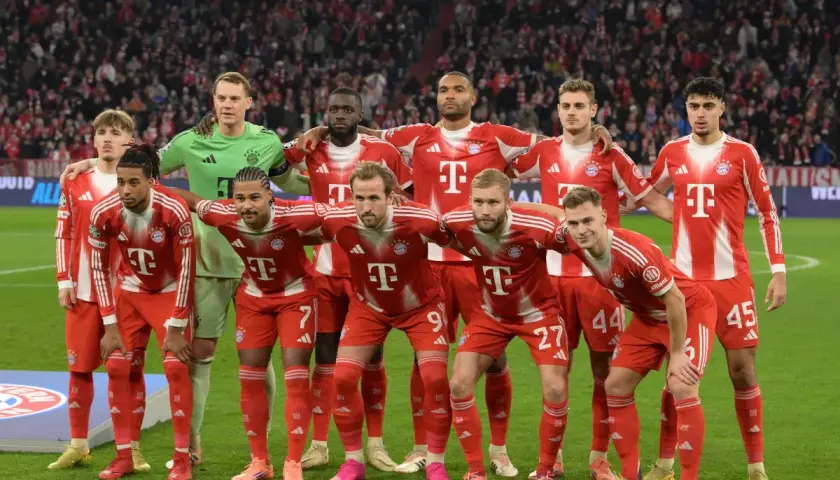 Drużyna Bayern Monachium – zawodnicy w czerwono-białych strojach pozują do zdjęcia na murawie stadionu, z trybunami pełnymi kibiców w tle.