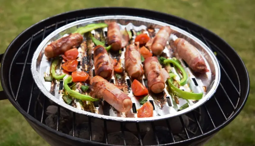 Pyszna kiełbasa na grilla, owinięta boczkiem, pieczona z papryką i pomidorkami na aluminiowej tacce.