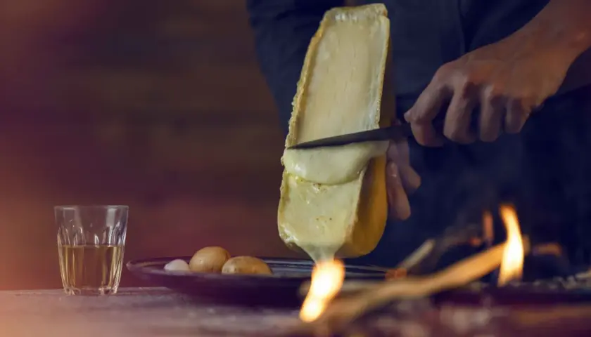 Un morceau de fromage à raclette fond sur un feu, rappelant son origine alpine. Des pommes de terre et un verre de vin complètent la scène.