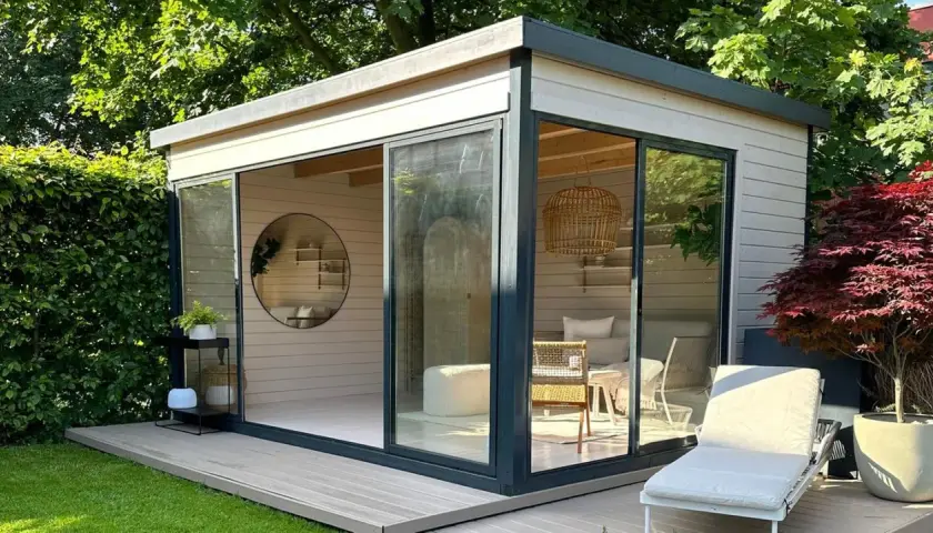 Modernes Gartenhaus mit Glasfront, das zum Streichen einlädt. Gemütliche Inneneinrichtung mit Rattan-Akzenten und Liegestuhl im Freien.