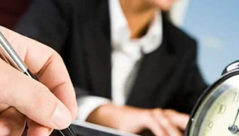 A hand holds a pen, poised to take notes, as a person in a suit works at a laptop. This scene evokes the process of creating board meeting minutes examples.