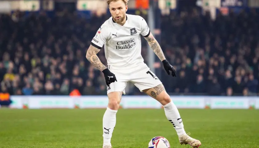 Piłkarz w białej koszulce z tatuażami na rękach i nogach, gdzie gra puchacz, przygotowuje się do strzału na zielonej murawie stadionu.