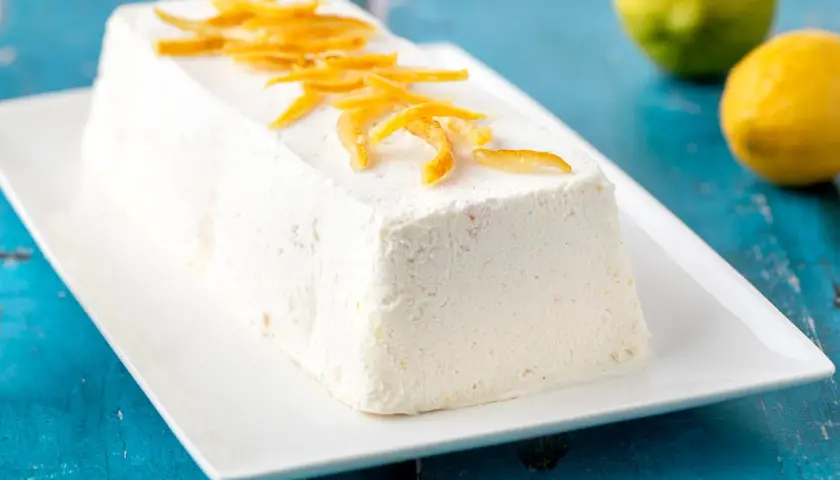 Delizioso semifreddo al limone, guarnito con scorzette candite, su un piatto bianco. Due limoni freschi sullo sfondo.