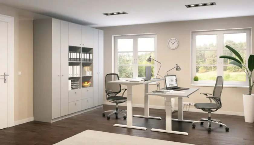 Ein helles, modernes Homeoffice mit zwei höhenverstellbaren Schreibtischen, Bürostühlen, einem großen Schrank und Fenstern mit Blick ins Grüne.