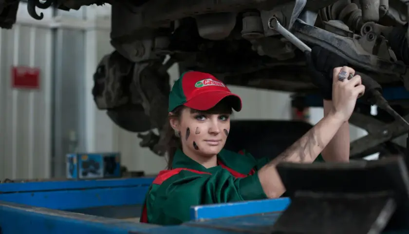 Mécanicienne sous une voiture, le visage barbouillé de cambouis. Elle est experte automobile, mais son regard suggère un abus de pouvoir.