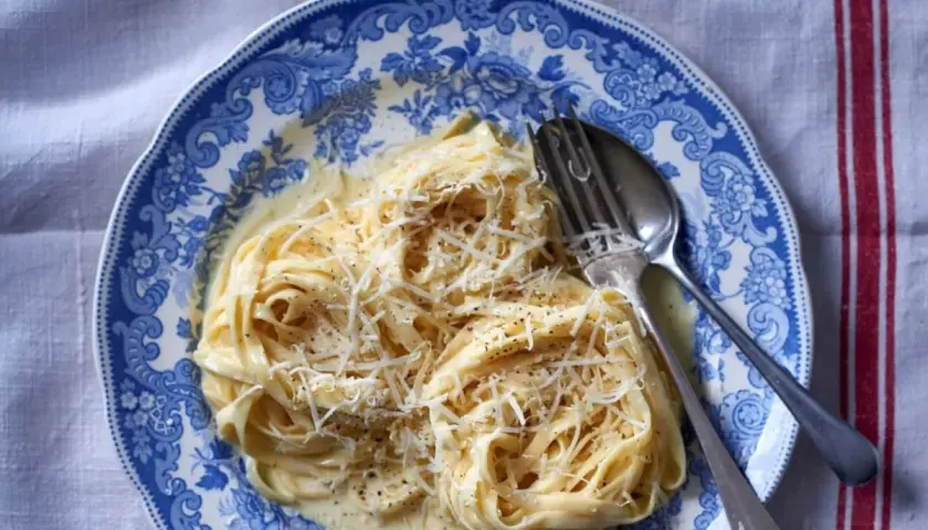 Tagliatelle w kremowym sosie alfredo, posypane parmezanem, na talerzu z niebieskimi wzorami.