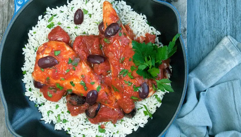 Un plat de poulet sauce tomate savoureux, servi sur un lit de riz blanc, garni d'olives noires et de persil frais.