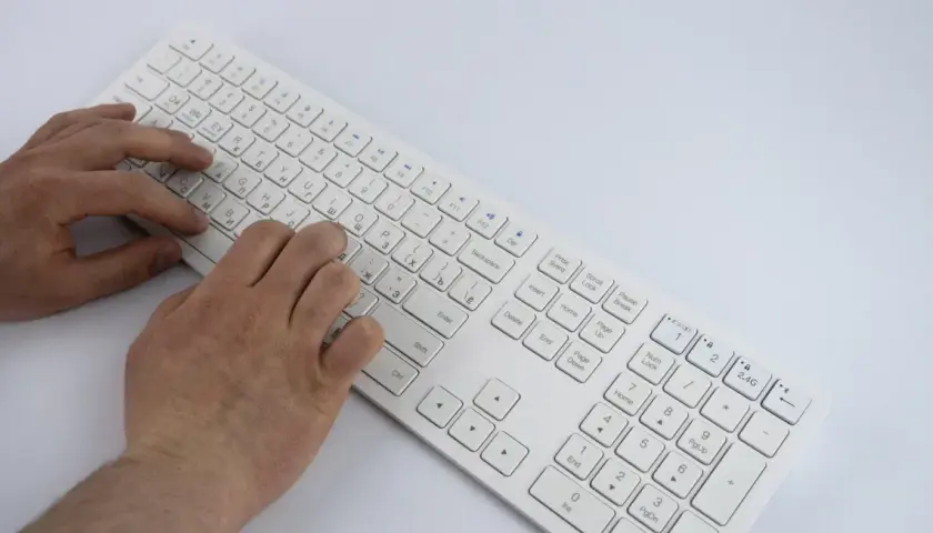 Dłonie piszą na białej klawiaturze laptopa. Widoczny jest opis klawiszy, w tym litery i symbole.