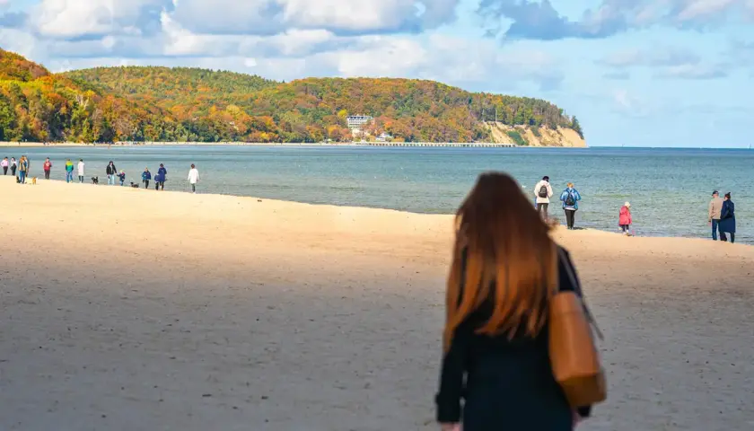 Jesień na nadmorskiej trasie spacerowej z Gdyni do Sopotu. Ludzie spacerują po piaszczystej plaży, podziwiając kolorowe drzewa na klifie.