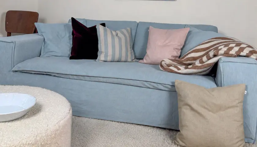 En ljusblå soffa med kuddar i olika färger och mönster. En mysig pläd ligger slängd över ena armstödet. Perfekt blå soffa inspiration för ett modernt hem.