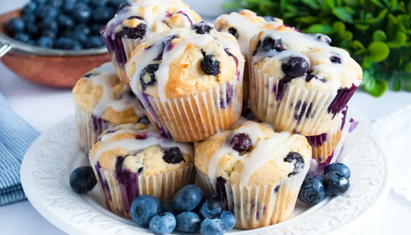 Stos świeżo upieczonych muffinek z borówkami, polanych lukrem, z kilkoma borówkami na talerzu.