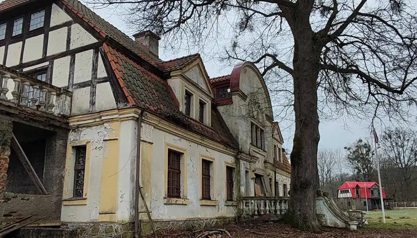 Zniszczony dworek w Paraszyne, z charakterystyczną architekturą i opadającą fasadą, otoczony drzewami.