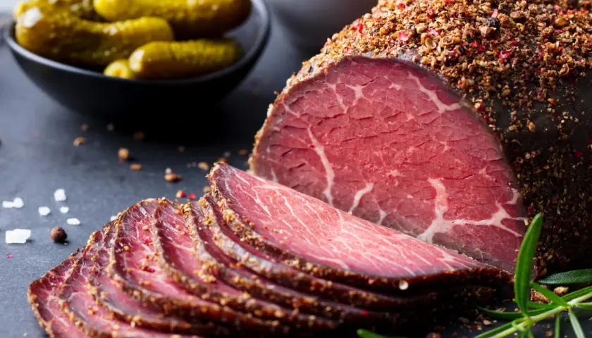 Pokrojone plastry pastrami, przyprawione pieprzem, obok ogórków konserwowych. Zastanawiasz się pastrami co to? To aromatyczne, peklowane i wędzone mięso wołowe.