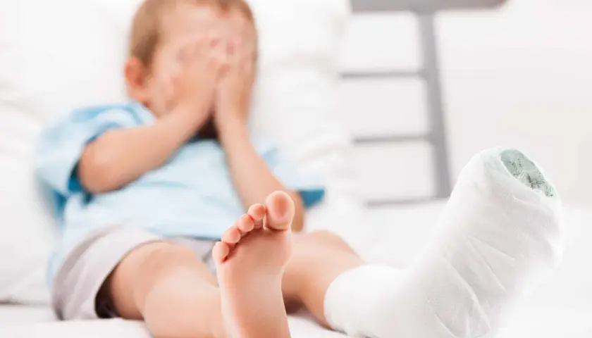 Chłopiec z zabandażowaną nogą, ukrywa twarz w dłoniach. Może to być skutek wypadku lub objaw krzywicy.