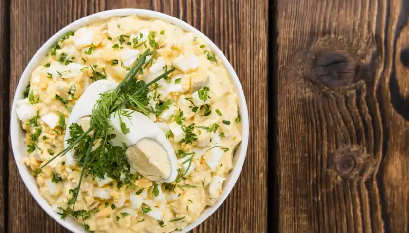 Insalata cremosa con uova di quaglia sode, erbe fresche e un uovo intero a metà come guarnizione, servita in una ciotola bianca su un tavolo di legno rustico.