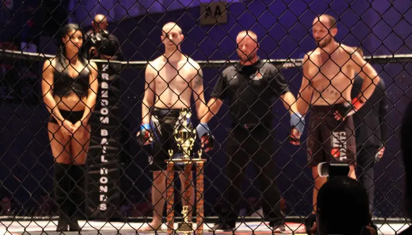 Ring girls i zawodnicy MMA czekają na rozpoczęcie freak fight. Trofeum lśni w blasku reflektorów.