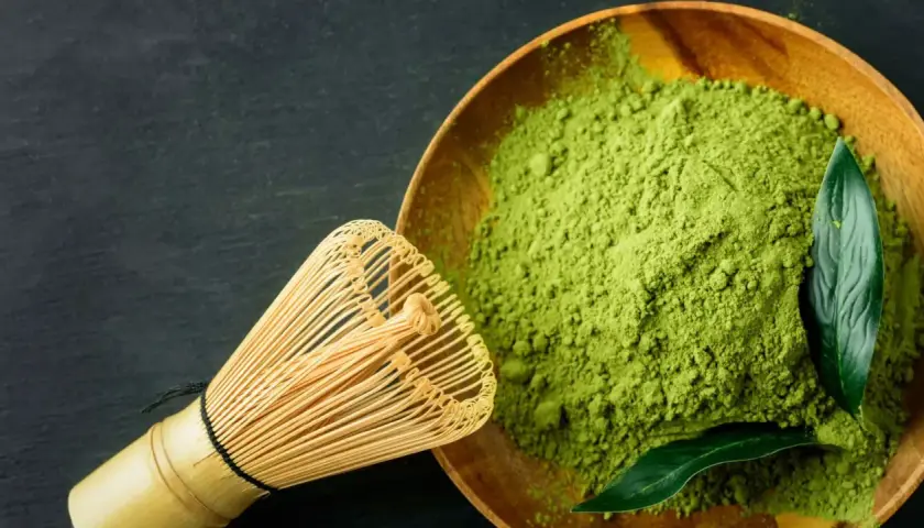 Ein hölzerner Löffel mit feinem, leuchtend grünem Matcha-Pulver und ein Bambusbesen daneben. Der Geschmack ist intensiv, leicht bitter.