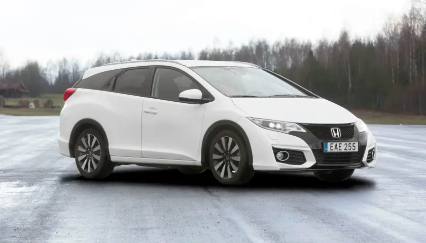 Vit Honda Civic Tourer parkerad på en fuktig väg med träd i bakgrunden.