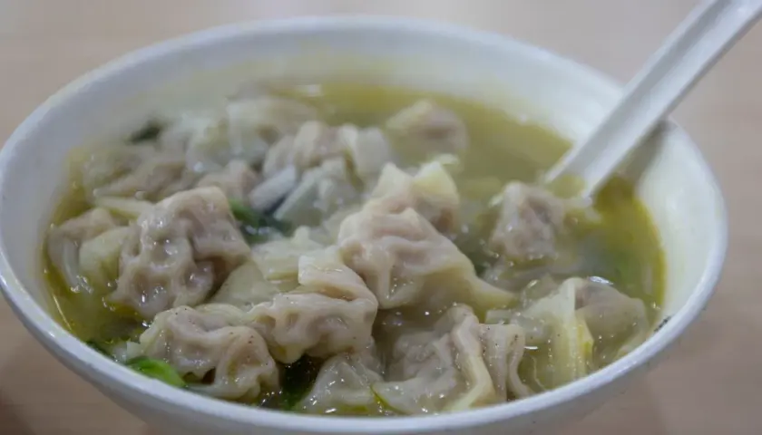 A miseczka pełna aromatycznej zupy wonton, z widocznymi pierożkami i zielonymi warzywami. Łyżka zanurzona w bulionie.