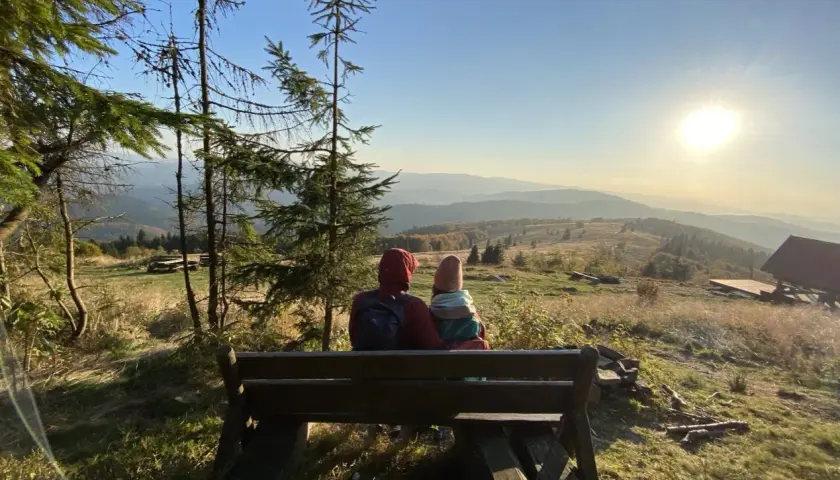 Para siedzi na ławce, podziwiając widok na góry. Słońce zachodzi nad horyzontem, malując niebo na złote i pomarańczowe barwy. To piękny widok z mogielicy z Jurkowa.