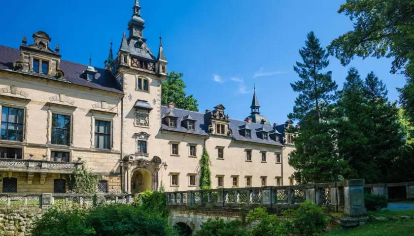 Elegancki hotel zamek z wieżą i mostkiem, otoczony zielenią, pod błękitnym niebem.