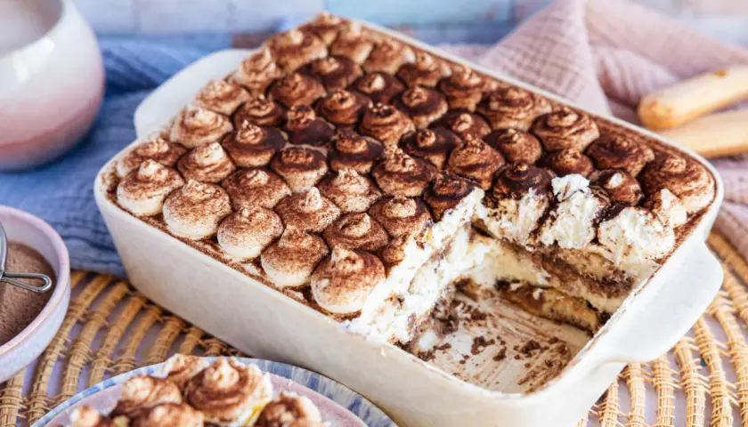 Cremiges, veganes Tiramisu ohne Alkohol, bestäubt mit Kakao. Ein Stück wurde bereits entnommen, was die Schichten aus Creme und Löffelbiskuits zeigt.