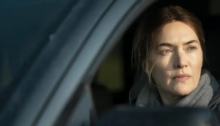 En kvinna med intensiv blick sitter i en bil, kanske på väg att lösa ett fall som i Mare of Easttown.