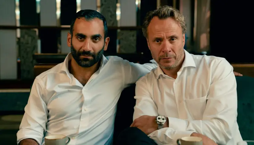 Två män i vita skjortor sitter vid ett bord med kaffe. En arm vilar på den andres axel. Scen från hundarna serie.