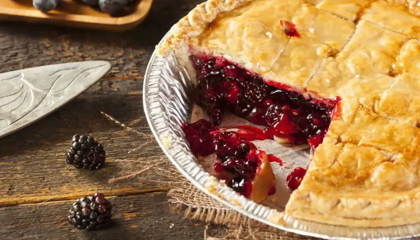 Una fetta di torta di more appena sfornata, con il ripieno succoso che cola. Due more fresche adagiate sul tavolo di legno.