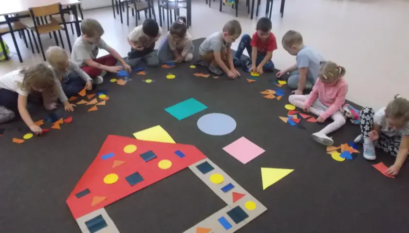 Dzieci w przedszkolu tworzą obrazki z kolorowych figur geometrycznych.