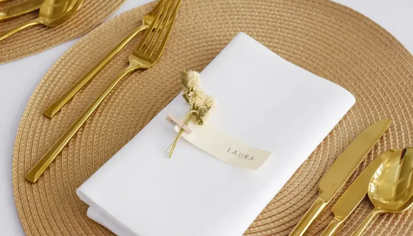 Elegante Tischdeko: Weiß gefaltete Servietten mit Namen "Laura", goldenes Besteck und kleine Trockenblumen auf einem geflochtenen Platzset.