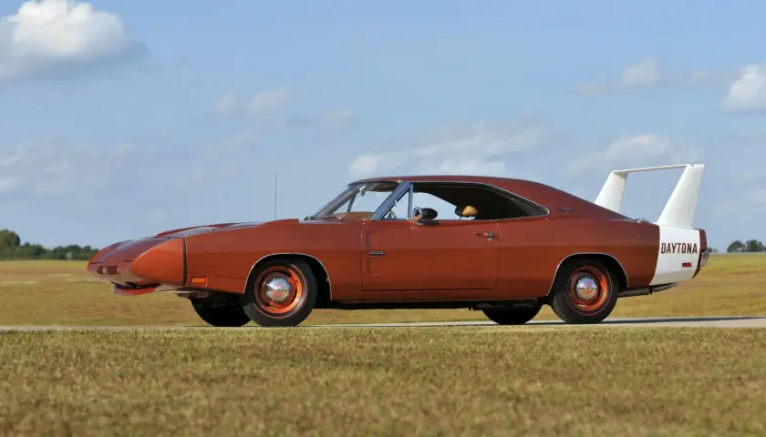 Pomarańczowy Dodge Daytona z charakterystycznym spojlerem i białym pasem z napisem "DAYTONA" na błotniku, zaparkowany na tle nieba z chmurami.