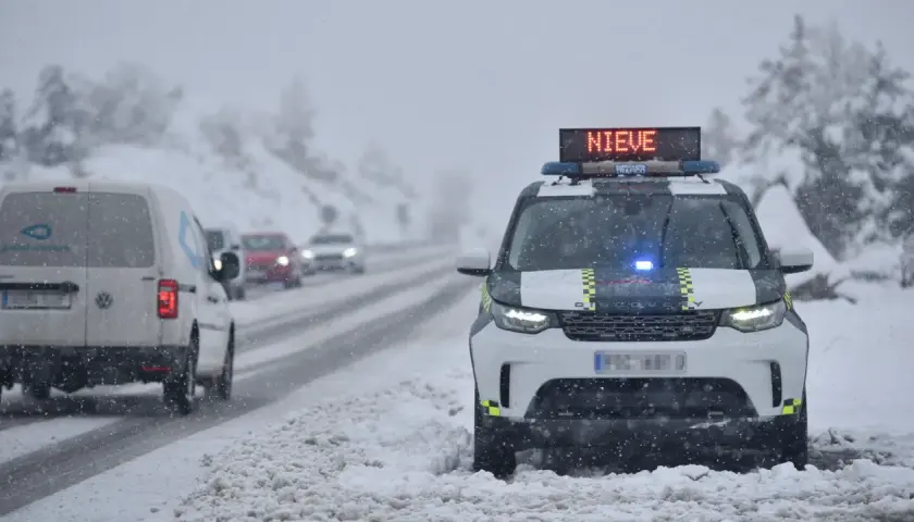 DGT alerta de nieve en carreteras. Un coche patrulla con luces azules y el mensaje "NIEVE" en su panel luminoso se detiene en un arcén nevado.