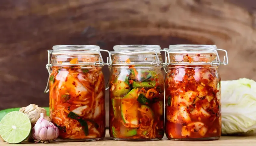 Drei Gläser mit koreanischer Vorspeise fermentierter scharfer Kohl (Kimchi) stehen bereit. Daneben liegen frische Zutaten wie Knoblauch, Ingwer und Limette.