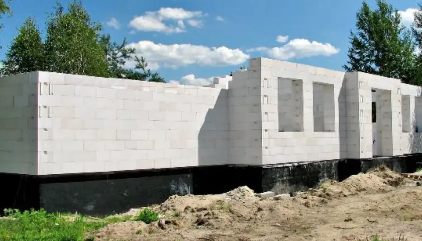 Nowe, białe ściany domu z bloczków betonowych, z otworami na okna i drzwi, stoją na czarnym fundamencie.