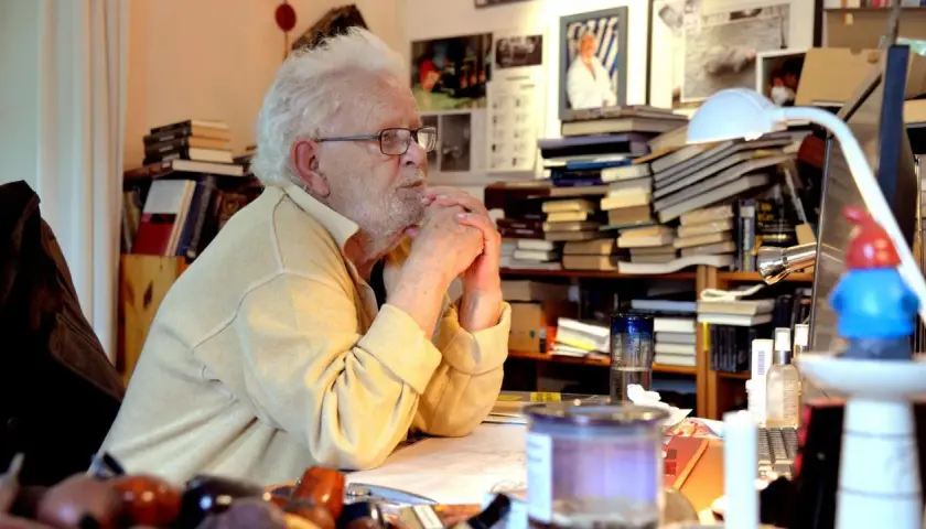 Älterer Mann mit Brille und weißem Haar sitzt am Schreibtisch, umgeben von Büchern.