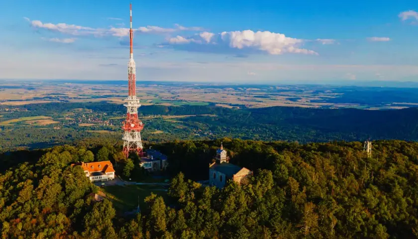 Wieża telekomunikacyjna na górze Ślęża góruje nad lasami i polami. Widać też budynki i kościółek.