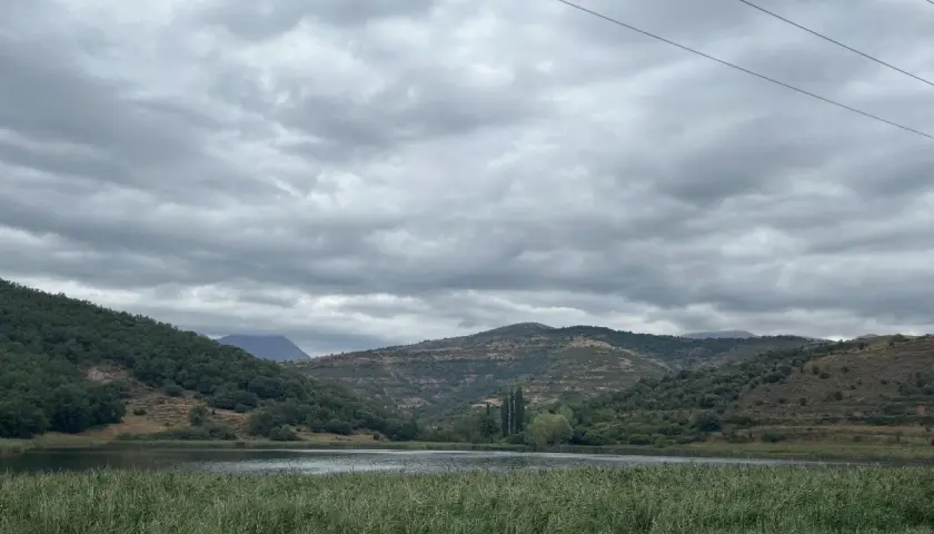 Paisaje sereno con un lago rodeado de colinas verdes y secas, bajo un cielo nublado. Las montañas a lo lejos parecen la geganta adormida.