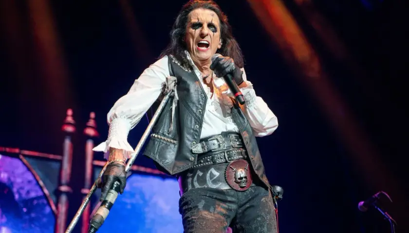 Alice Cooper na scenie z mikrofonem i kulami, w charakterystycznym makijażu i stroju.