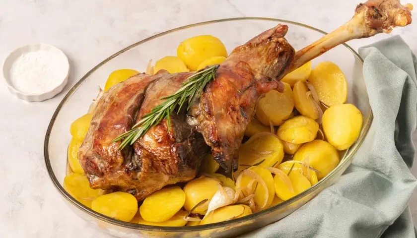 Pierna de cordero al horno dorada, servida con patatas asadas, romero y ajo en una fuente de vidrio.